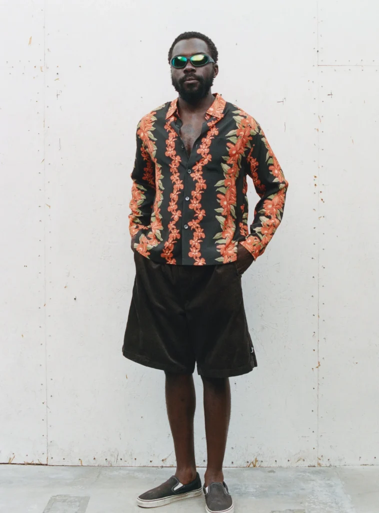 Person wear reflective sunglass, a black floral shirt with orange flower, black shorts, and dark slip-on shoe