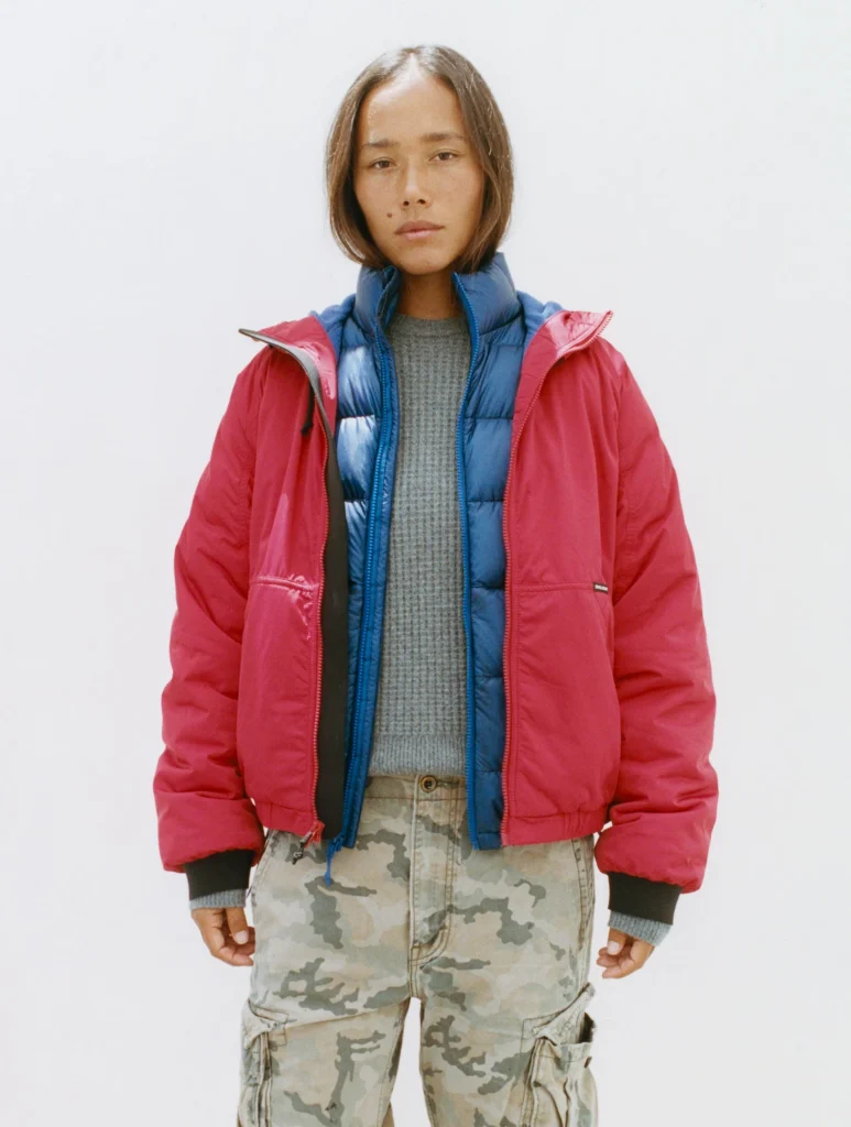 Person wear a red jacket layered over a blue puffer vest and gray sweater with camouflage cargo pant