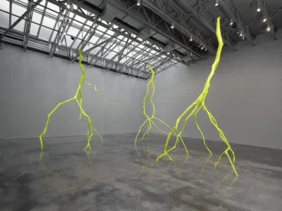 Three neon yellow-green, root-like branching sculptures rise across a minimalist gallery with polished concrete floors, gray walls, and an exposed industrial skylight ceil
