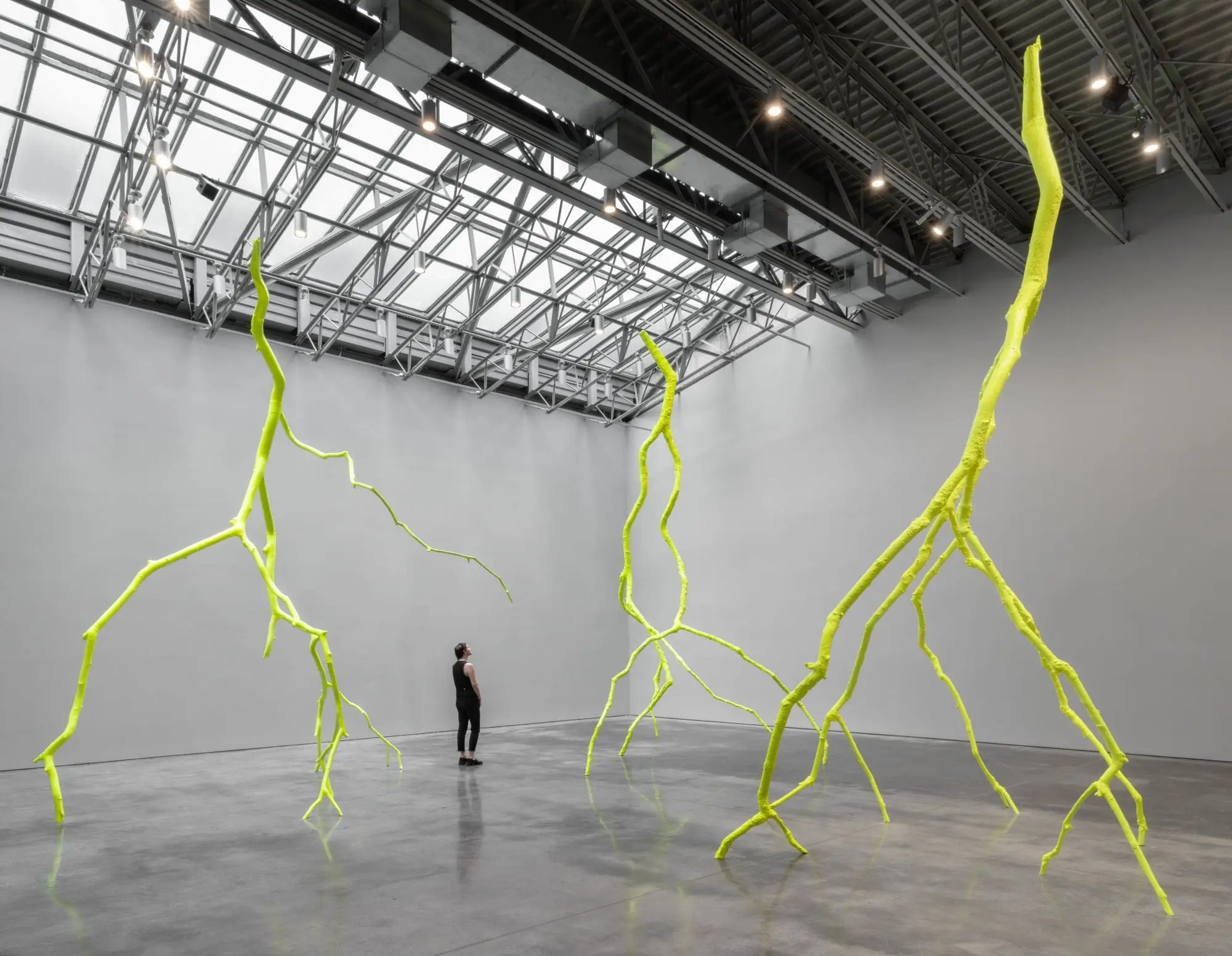 Three towering neon yellow-green, root-like branching sculptures fill a spacious contemporary gallery as a visitor stands between them beneath an industrial glass skylight.