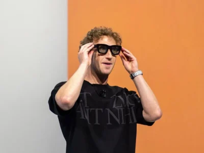 Mark Zuckerberg wear oversize black smart glasses onstage, adjust the frame with both hands against a bright orange and white background
