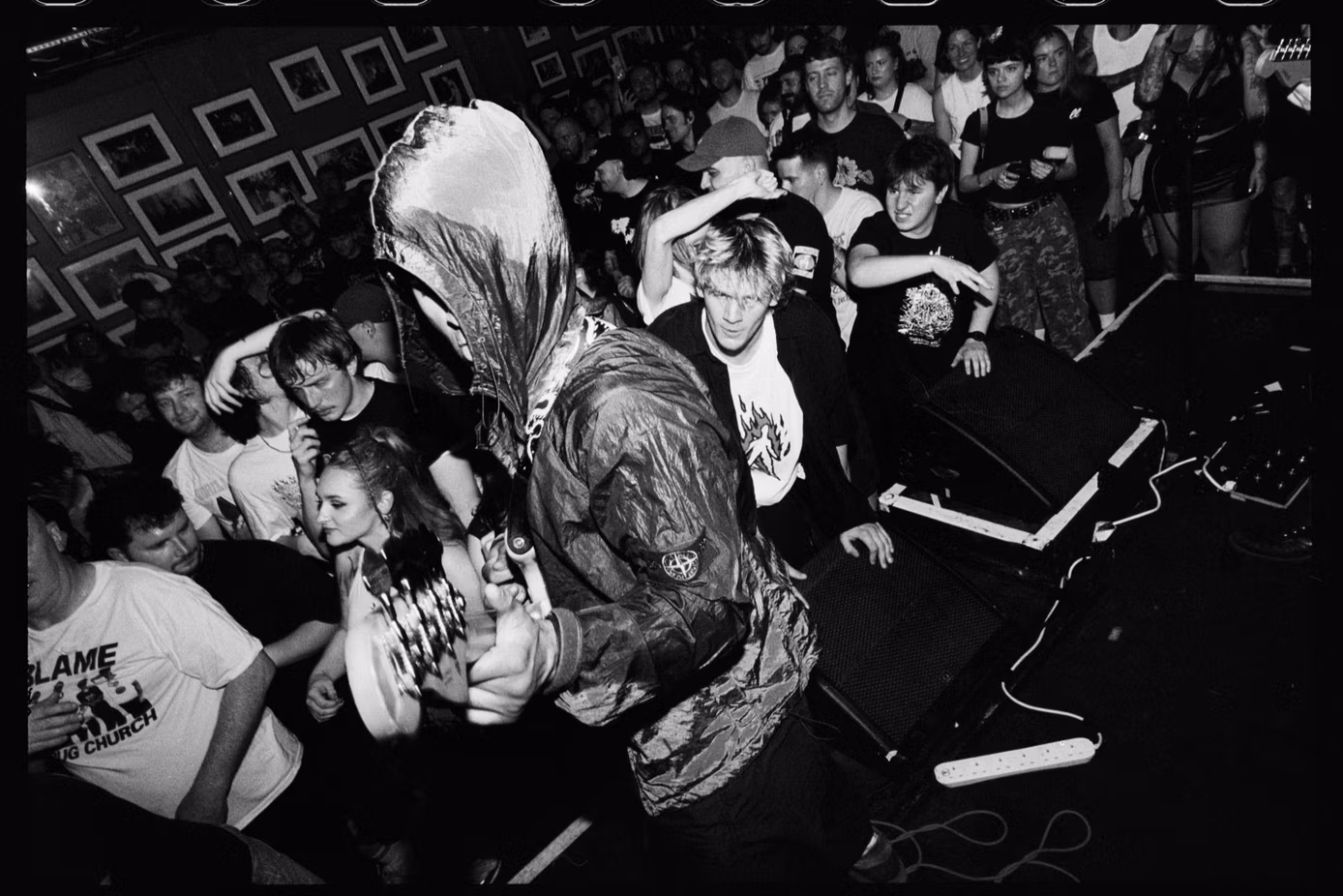 Black-and-white photograph of a packed hardcore show, with a vocalist in a hooded Stone Island jacket performing amid an energetic crowd beside the stage.