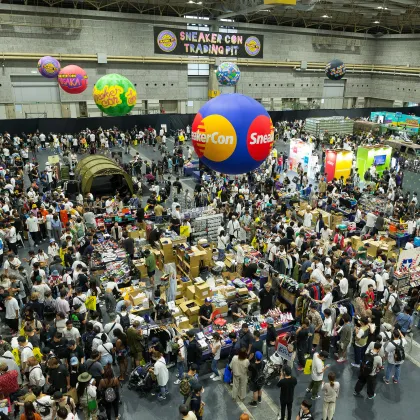 Overhead view of a packed Sneaker Con Trading Pit with crowds browsing vendor tables beneath colorful suspended Sneaker Con balloons.