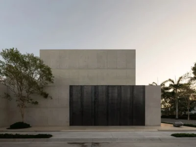 Symmetrical front façade of a minimalist concrete residence with towering black pivot doors, carefully placed landscaping, and warm evening light accentuating the architecture