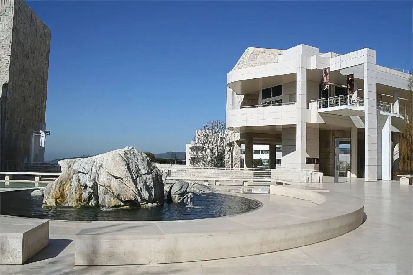 Modern stone plaza with a sculptural rock fountain in front of a contemporary white building under a clear blue sky.