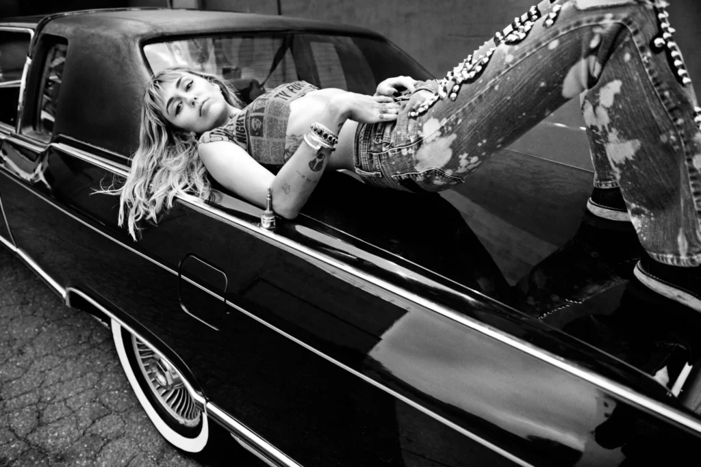 Miley Cyrus reclines across a vintage black car, wearing a newspaper-print crop top and studded jeans