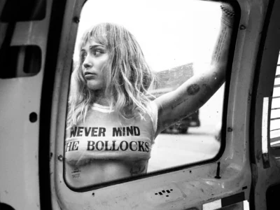 Black-and-white portrait of a tattooed Mylie Cyrus in a cropped “Never Mind the Bollocks” shirt, framed through a vehicle window