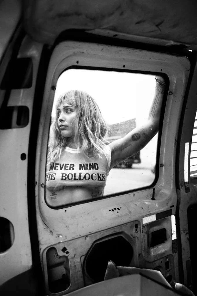 Black-and-white portrait of a tattooed Mylie Cyrus in a cropped “Never Mind the Bollocks” shirt, framed through a vehicle window