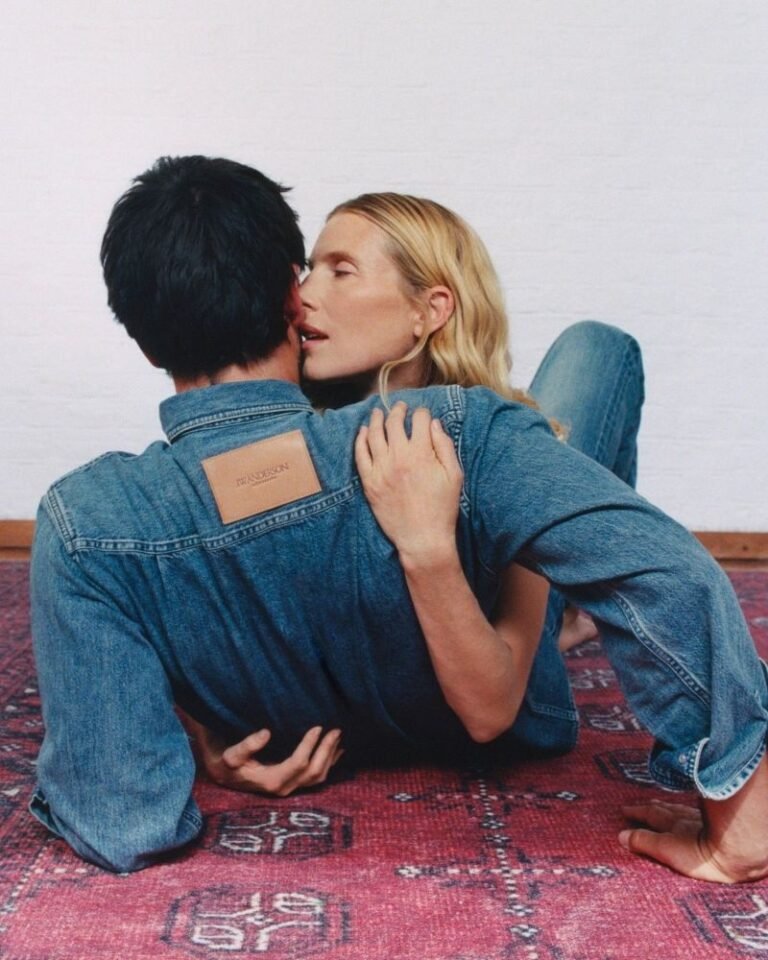 Two people in blue denim embrace and kiss while kneeling on a patterned red rug against a white wall