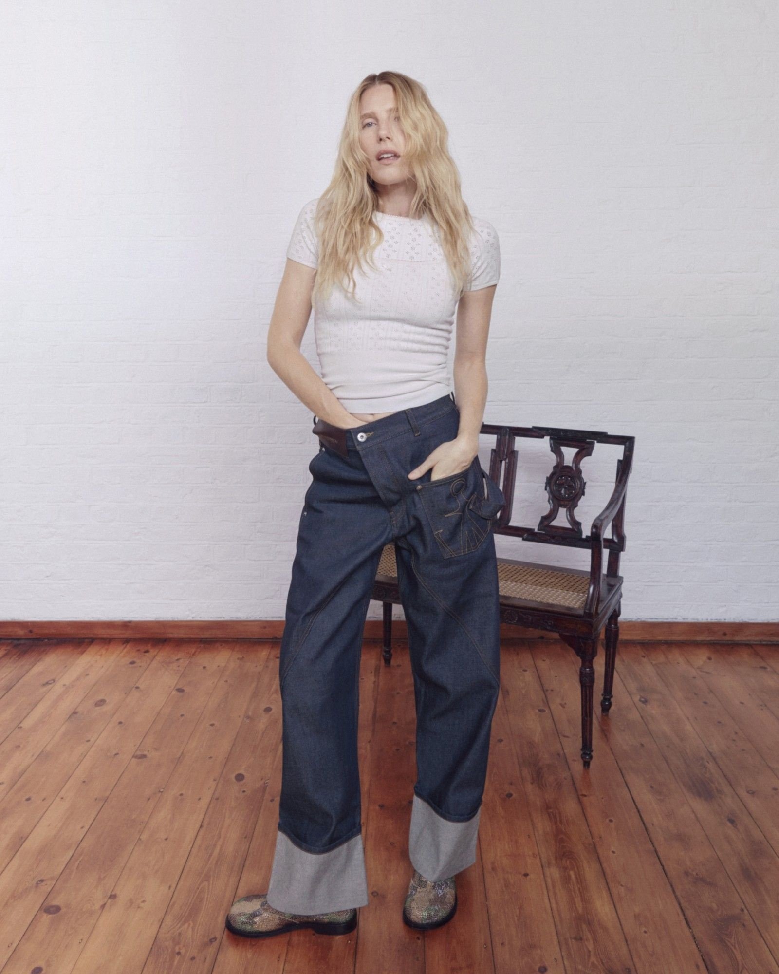 Blonde model stands in a white knit top and layered dark, wide-leg denim trousers beside an ornate wooden chair