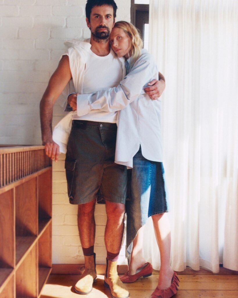 A man in a white sleeveless shirt and dark shorts embraces a woman in a pale blue shirt and denim skirt indoors