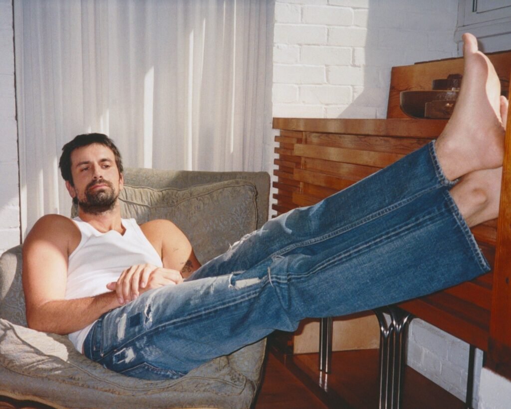 Bearded man reclining in a chair, wearing a white tank top and distressed blue jeans with legs raised on a table