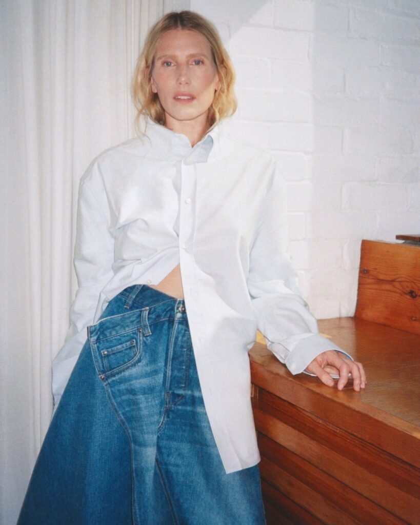 Blonde woman in a white shirt and asymmetrical blue denim skirt leans against a wooden dresser in a white room