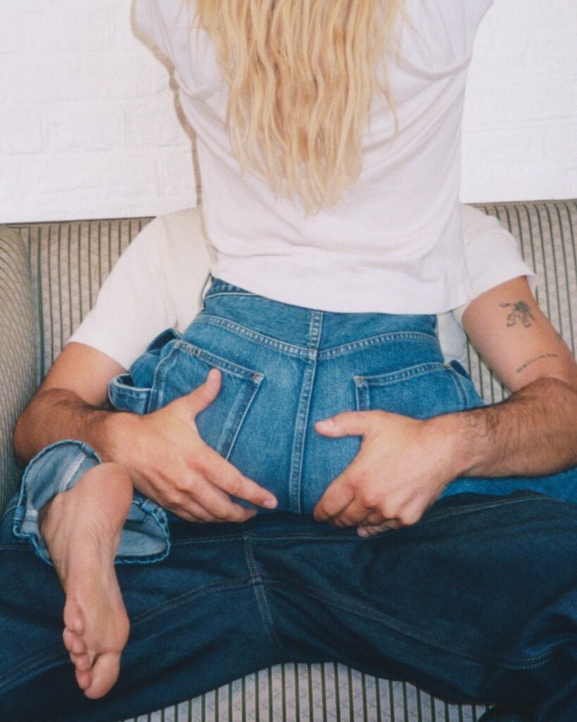 Cropped couple seated on a striped sofa, embracing a large blue denim garment; one wears a white shirt and has tattoos
