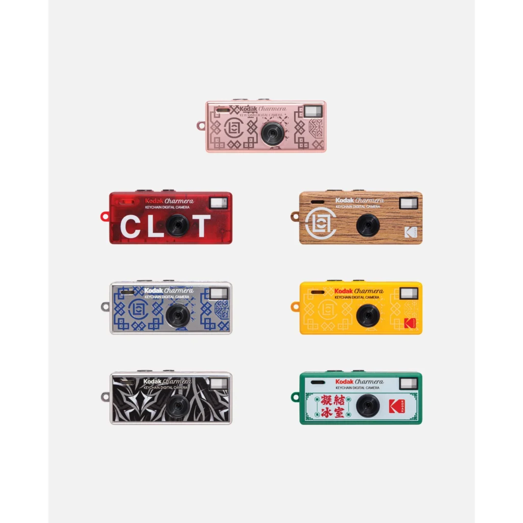 Seven Kodak Charmera keychain digital cameras in pink, red, woodgrain, yellow, blue, black, and green designs arranged on