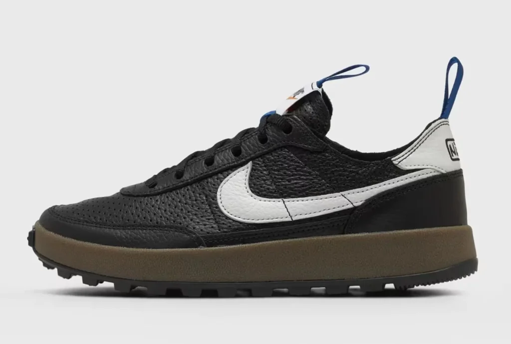Black low-top sneaker with white swoosh, brown midsole, rugged black outsole, and blue heel and tongue pull tabs on a white