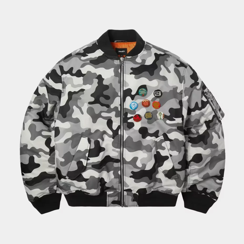 Gray, black, and white camouflage HUF bomber jacket with orange lining, black trim, and colorful pins on the chest against a