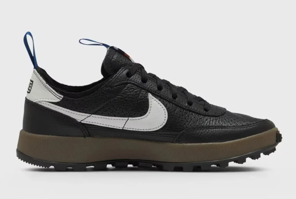 Side view of a black leather sneaker with a white swoosh, brown midsole, rugged black outsole, and blue pull tabs on a white