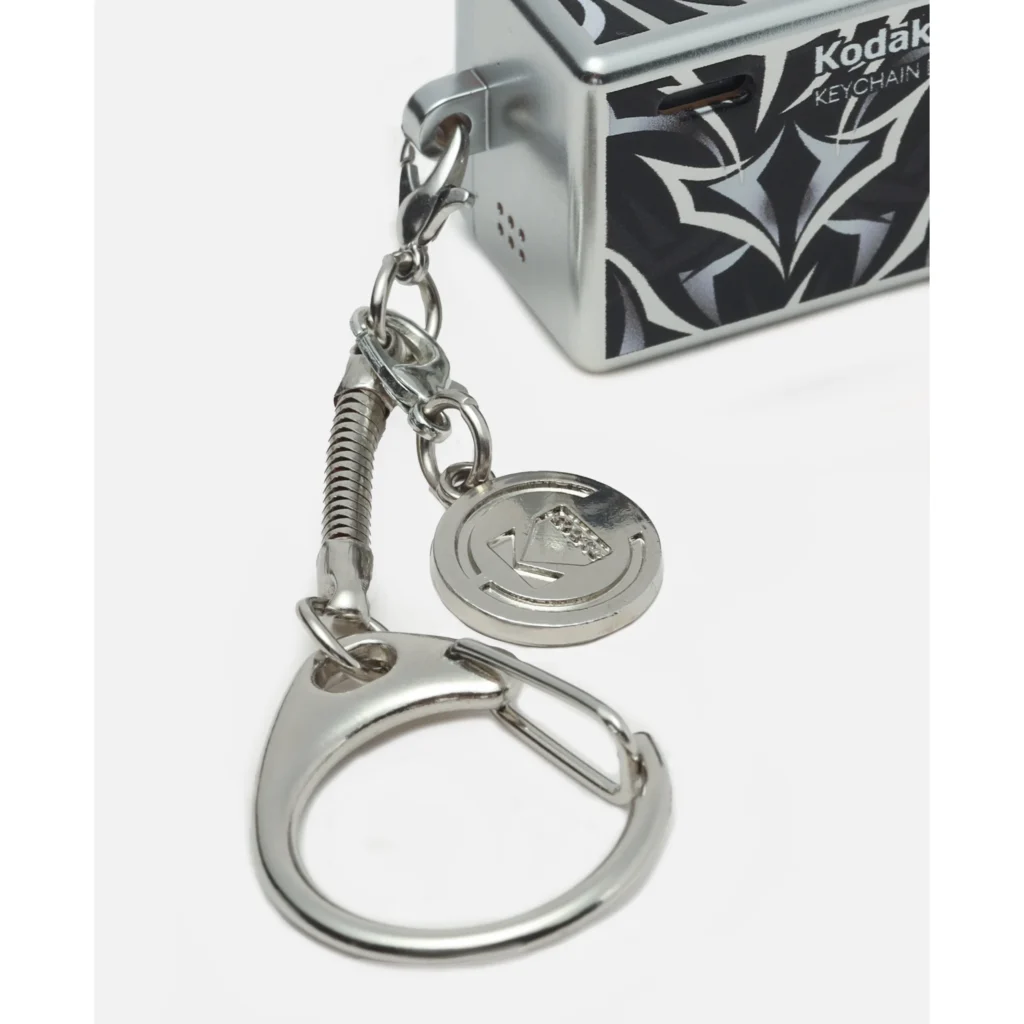Silver metal keychain with a carabiner, coiled charm, round medallion, and patterned Kodak box in the background