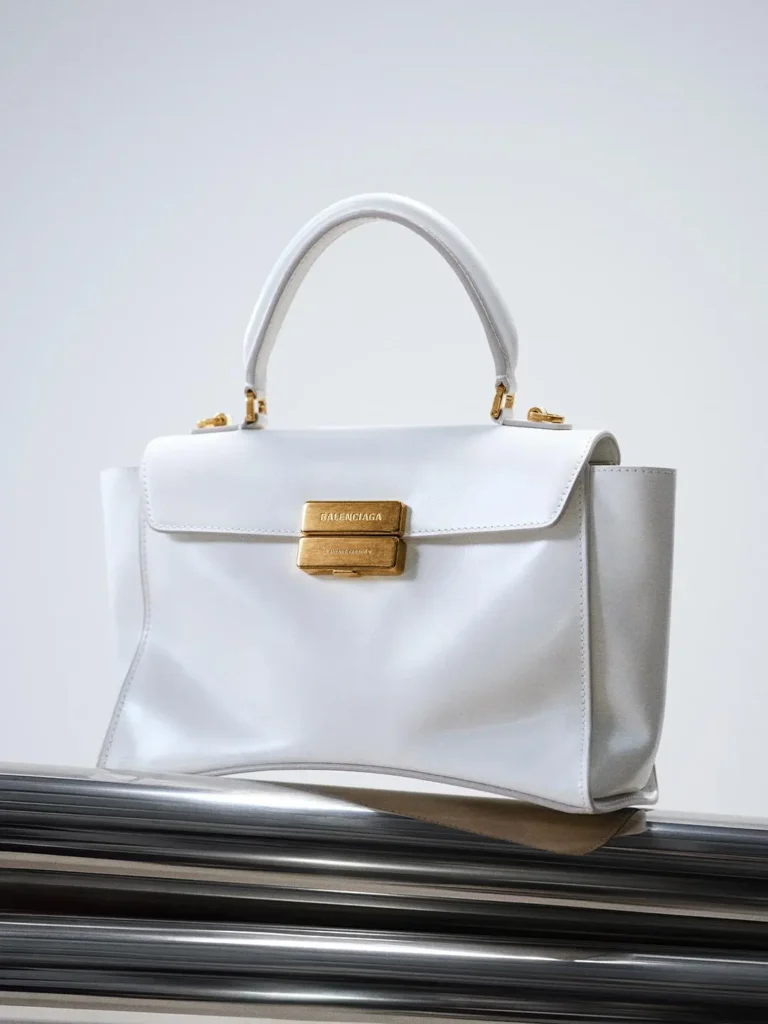 White leather Balenciaga handbag with a top handle and gold clasp, displayed on polished black molding