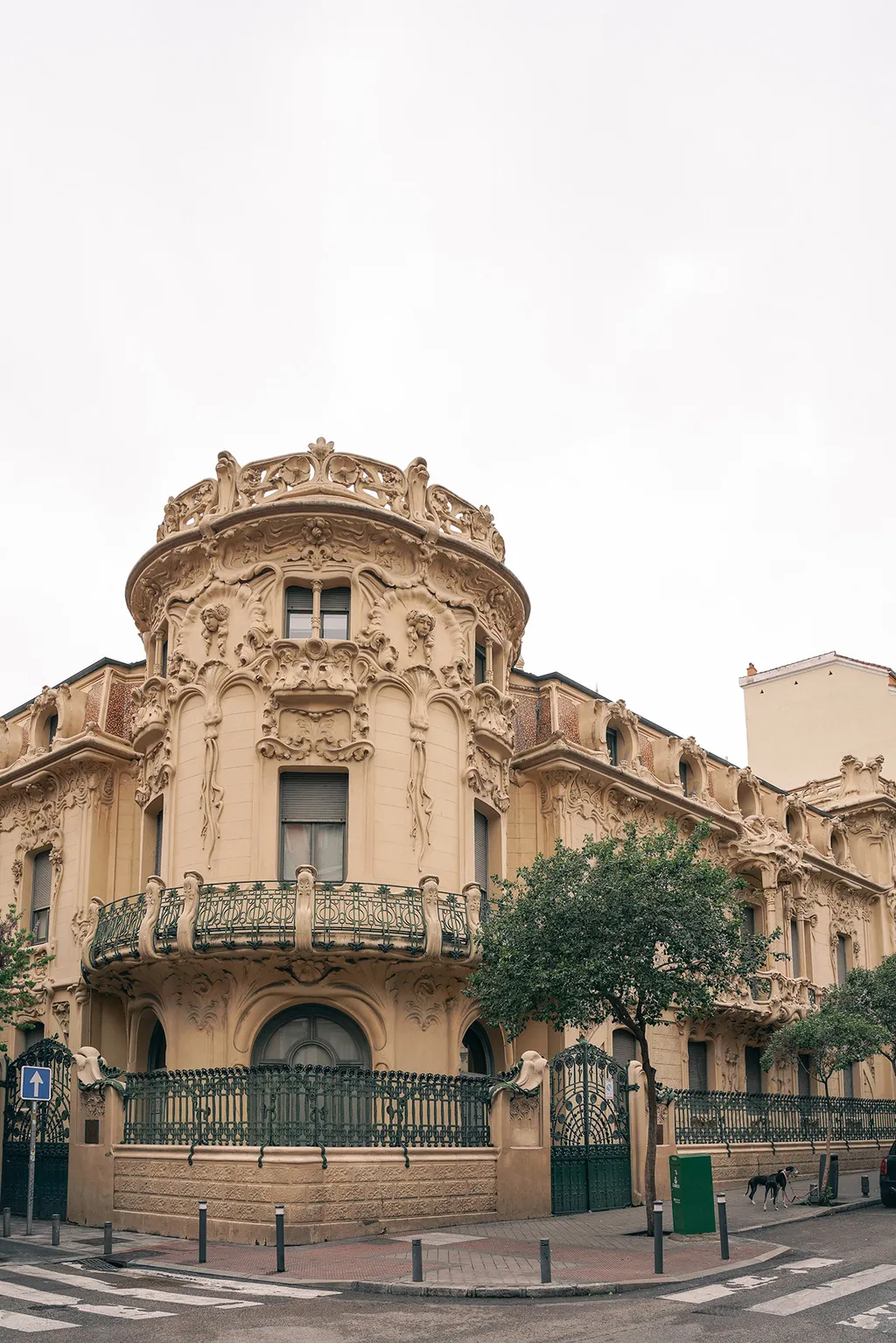 Ornate cream-colored Art Nouveau building with sculpted façade, curved balcony, iron gates, trees, and a street corner