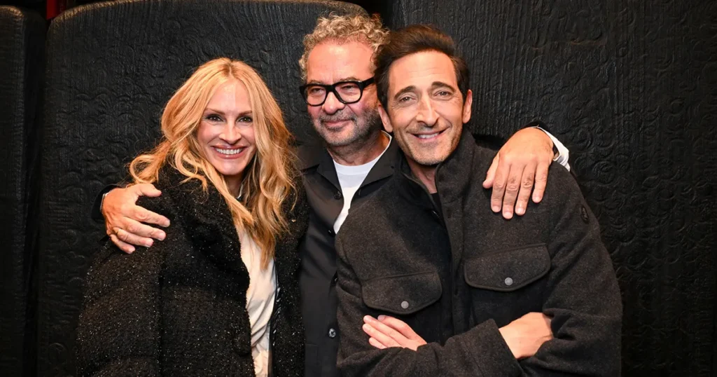 Left Julia Roberts, Mid Remo Luffini, Right Adrian Brody pose together against a black textured backdrop; the woman wears a sparkly black coat and the men dark
