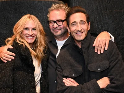 Left Julia Roberts, Mid Remo Luffini, Right Adrian Brody pose together against a black textured backdrop; the woman wears a sparkly black coat and the men dark
