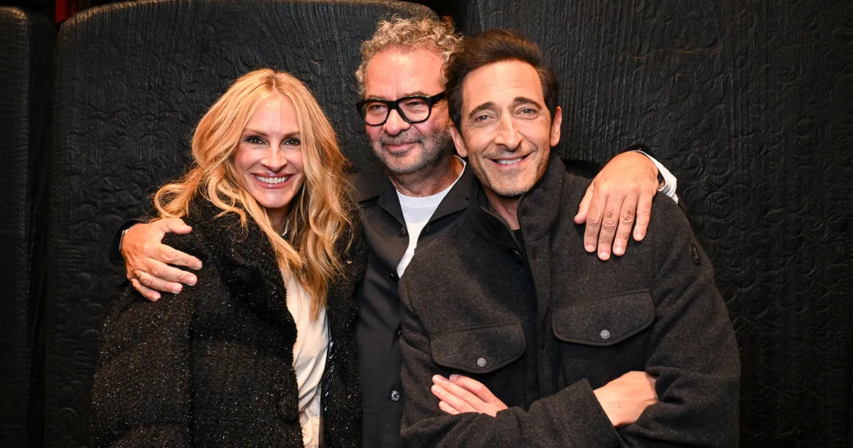 Left Julia Roberts, Mid Remo Luffini, Right Adrian Brody pose together against a black textured backdrop; the woman wears a sparkly black coat and the men dark