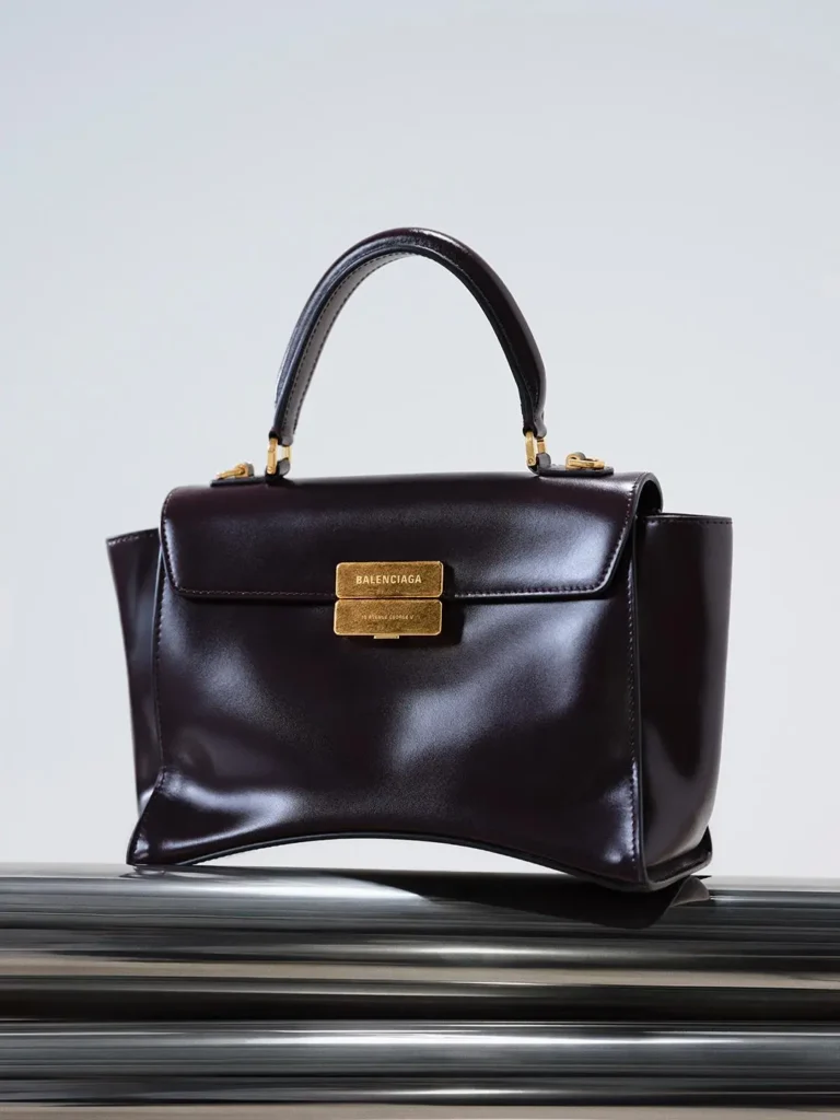 Dark burgundy leather Balenciaga handbag with top handle and gold clasp, displayed on stacked black surfaces against a pale