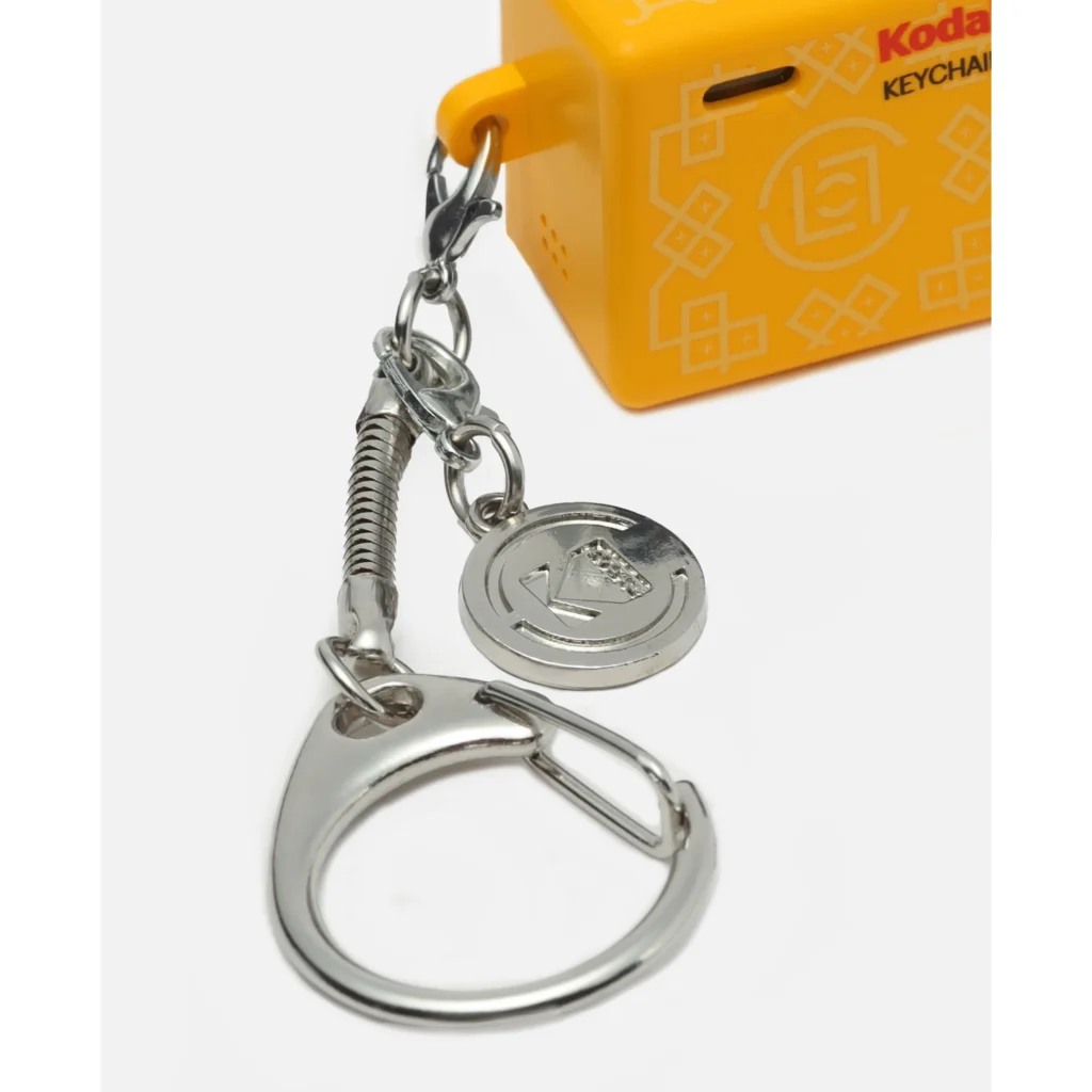 Close-up of a yellow Kodak camera keychain with silver carabiner, chain, and circular charm on a white background