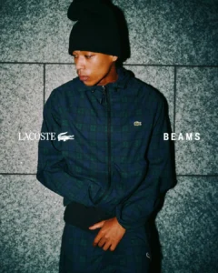 Person in a black beanie and navy-green plaid Lacoste jacket, standing against a gray tiled wall