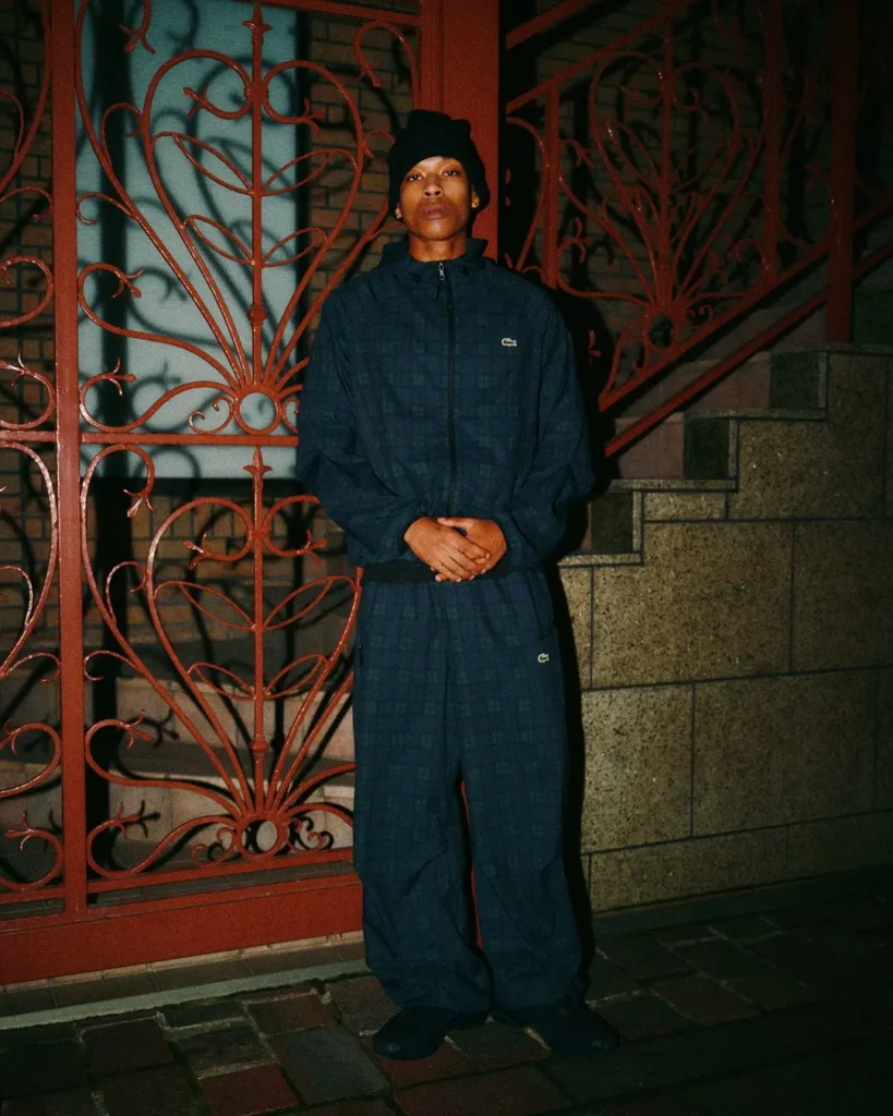 Person in a navy plaid tracksuit and black beanie stands with hands clasped before an ornate red iron gate and stone wall