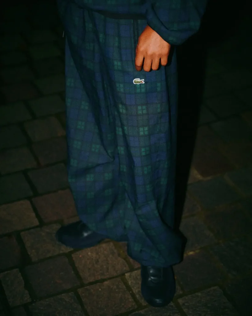 Close-up of a person wearing blue-green plaid pants with a crocodile logo and black shoes on a cobblestone pavement