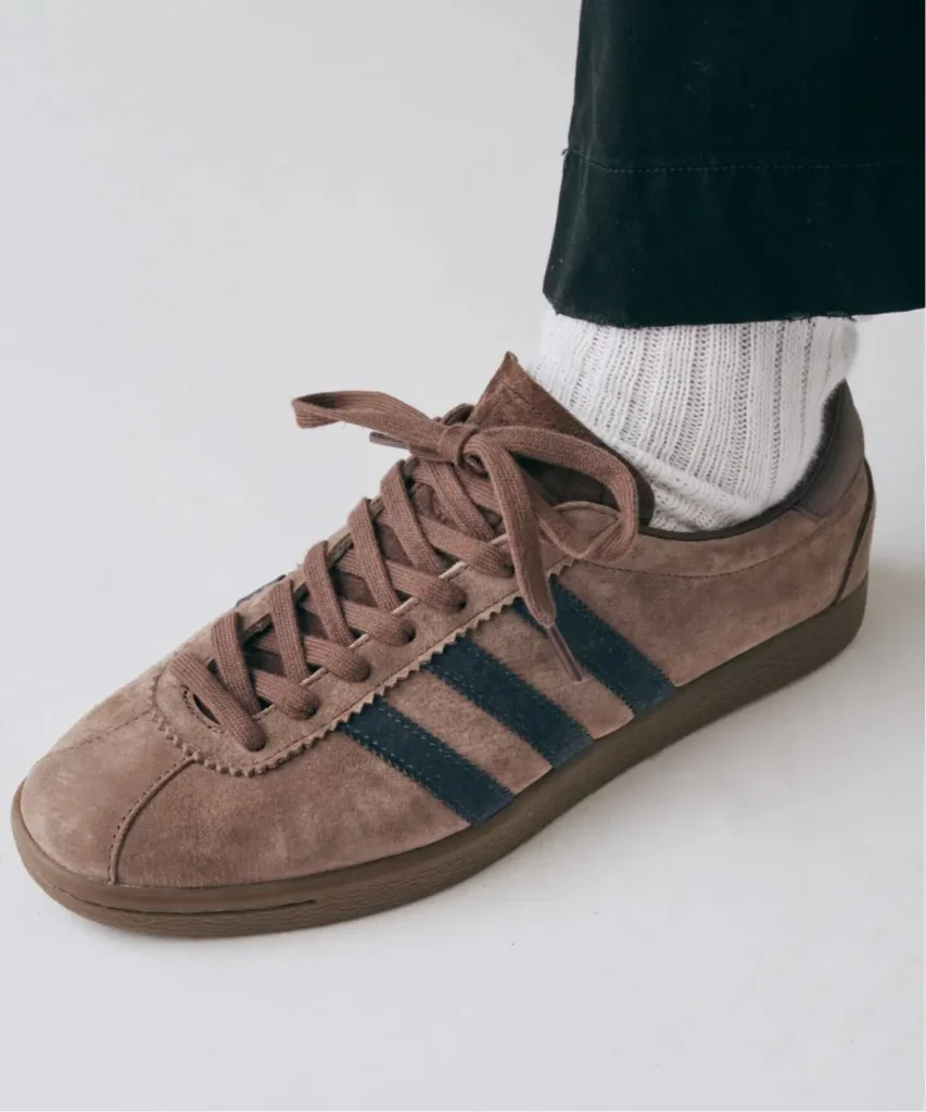 Close-up of a brown suede adidas sneaker with black stripes, worn with a white ribbed sock against a white background