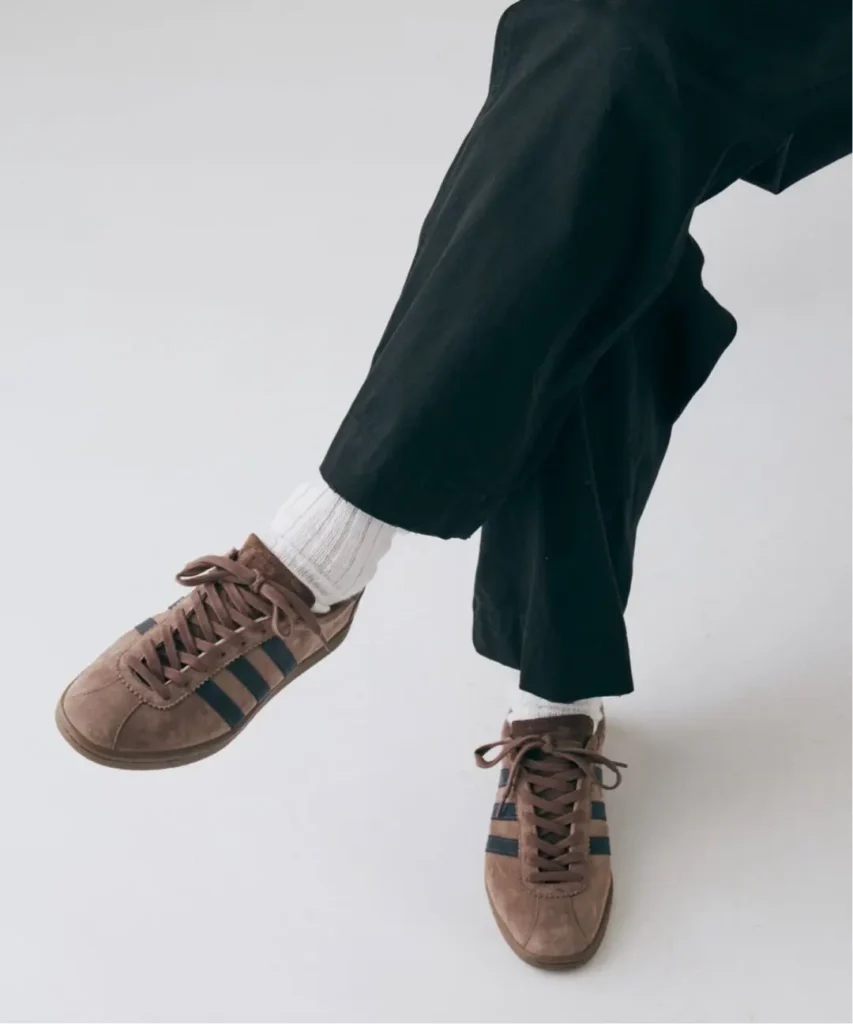 Close-up of brown suede adidas sneakers with dark stripes, white socks, and black cropped trousers against a pale background