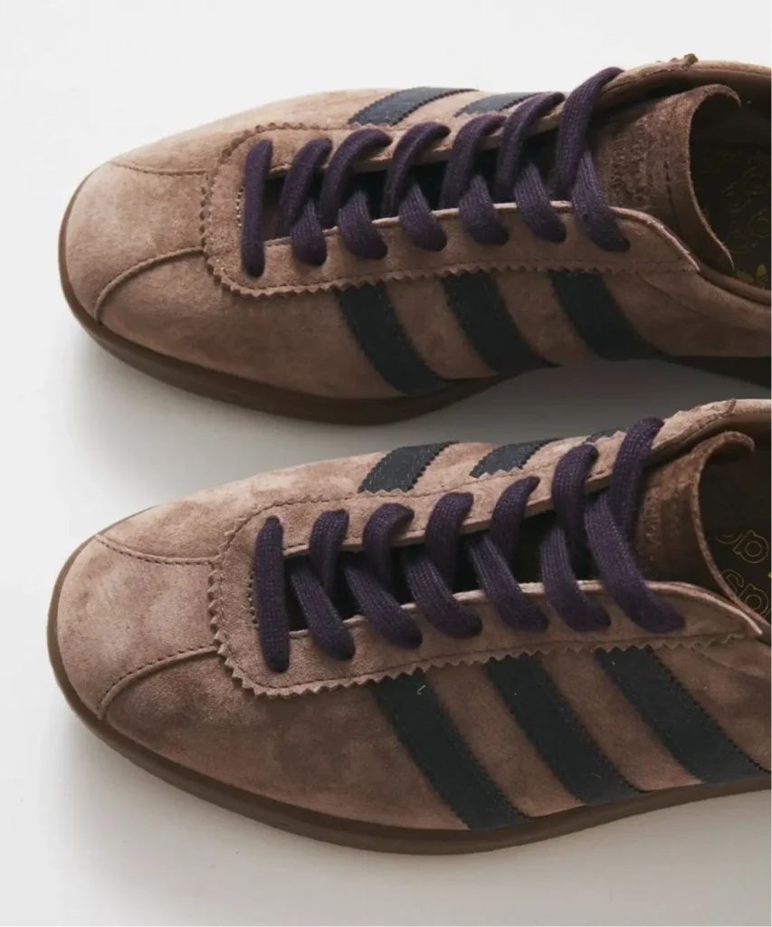 Two brown suede adidas sneakers with dark purple laces and black stripes on a white background