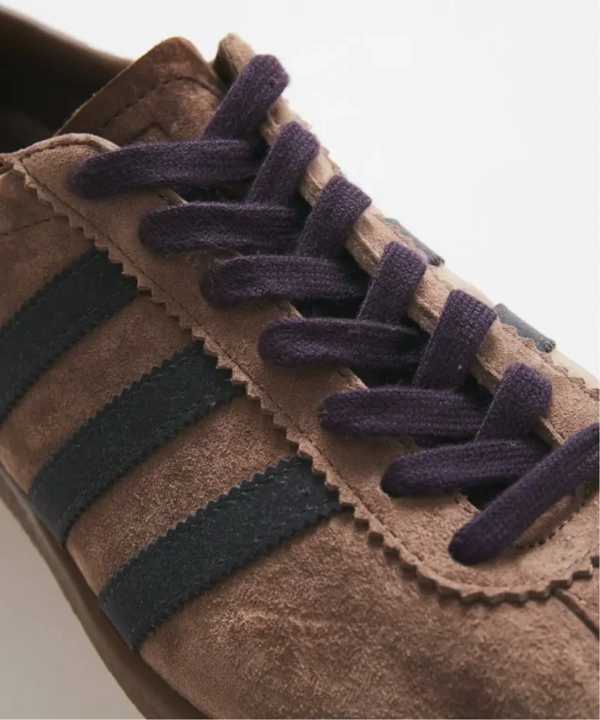 Close-up of a brown suede adidas Tobacco sneaker with black stripes and dark purple laces against a light background