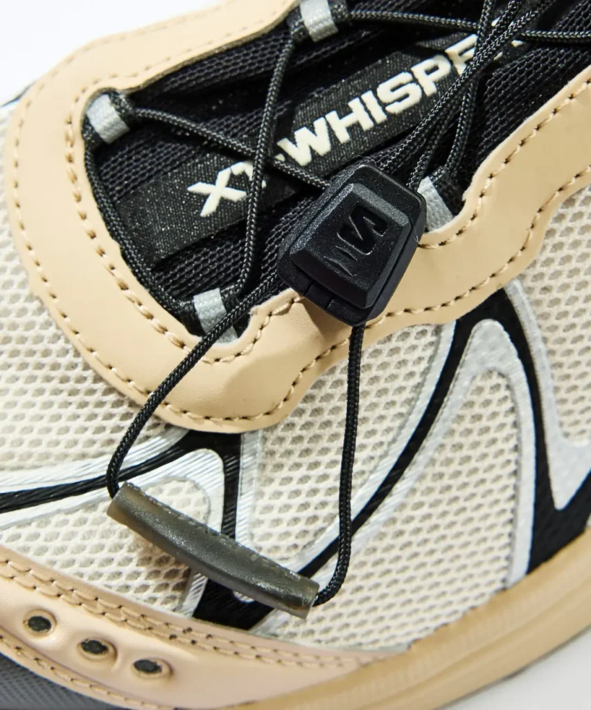 Close-up of a tan and black sneaker with mesh panels, black laces, and a raised lace toggle over a patterned sole