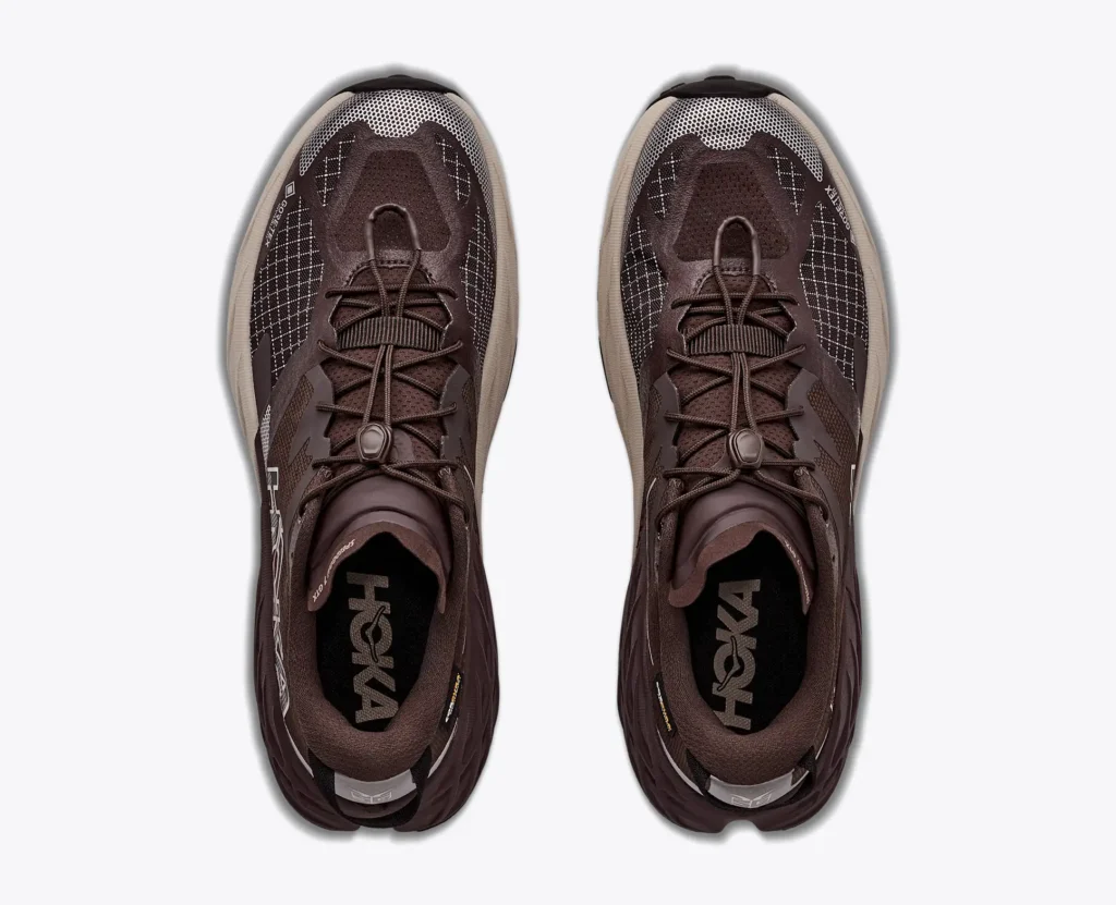 Top-down view of a pair of dark brown HOKA trail shoe with mesh uppers, laces, and thick textured soles on a white