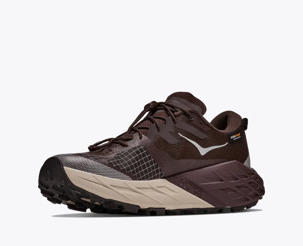 Brown HOKA trail shoe with beige midsole, black lugged outsole, silver logo and Cordura label on a white background