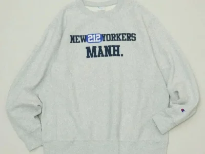 Light gray crewneck sweatshirt with navy “NEW YORKERS MANH.” lettering and a small Champion logo on the sleeve, laid flat