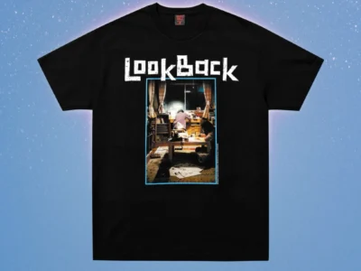 Black T-shirt with white “Look Back” lettering and a framed indoor scene graphic, displayed on a blue speckled background