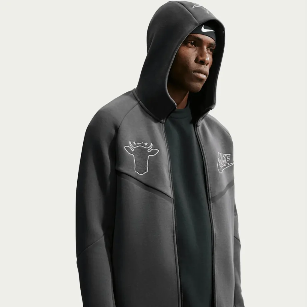 Model in a dark gray Nike hooded jacket with a white cow-head chest graphic, standing against a pale background