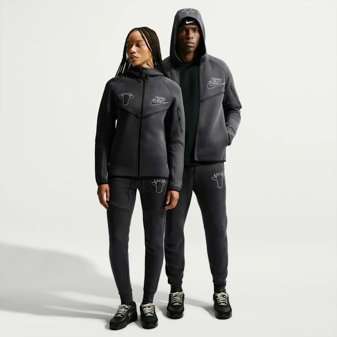 Two models wear black Nike tracksuits with cow-head graphics and sneakers against a plain light background