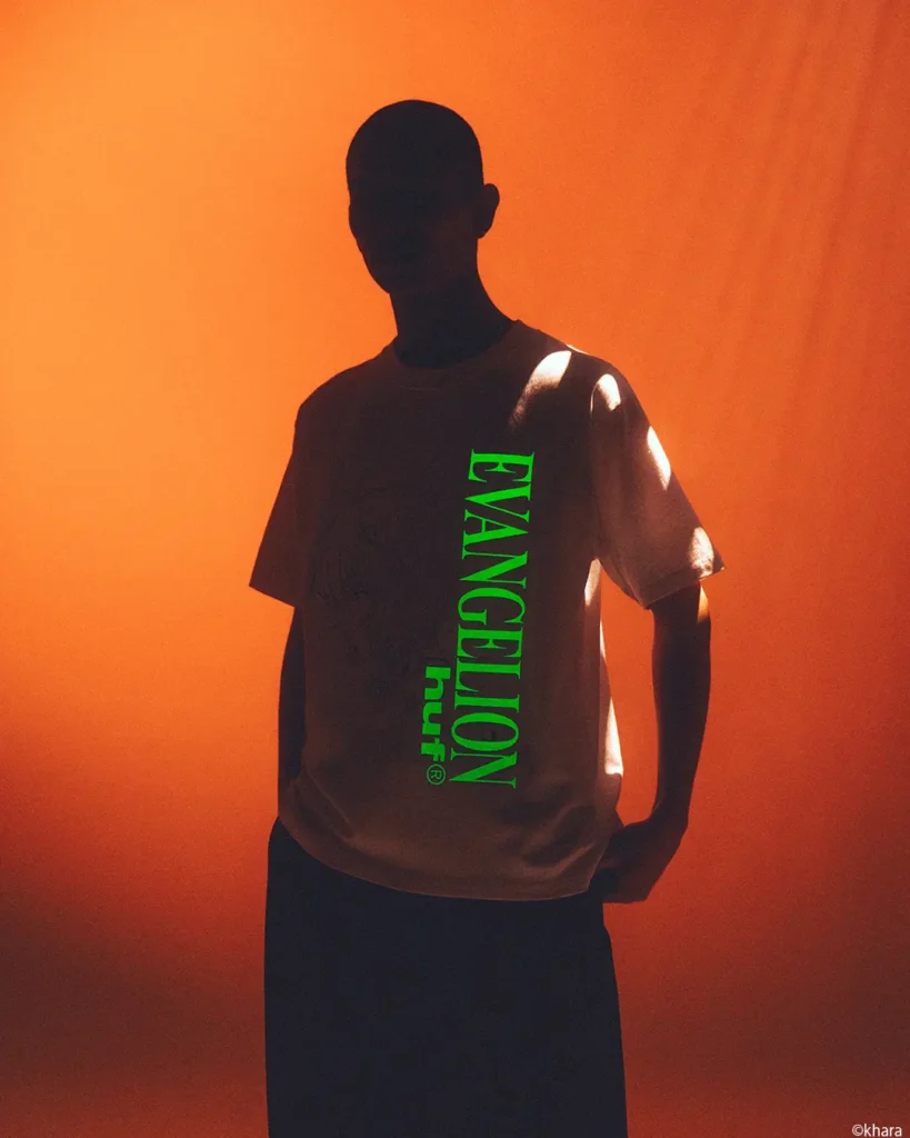 Silhouetted person in a brown T-shirt with vertical neon-green “EVANGELION” text, against an orange background