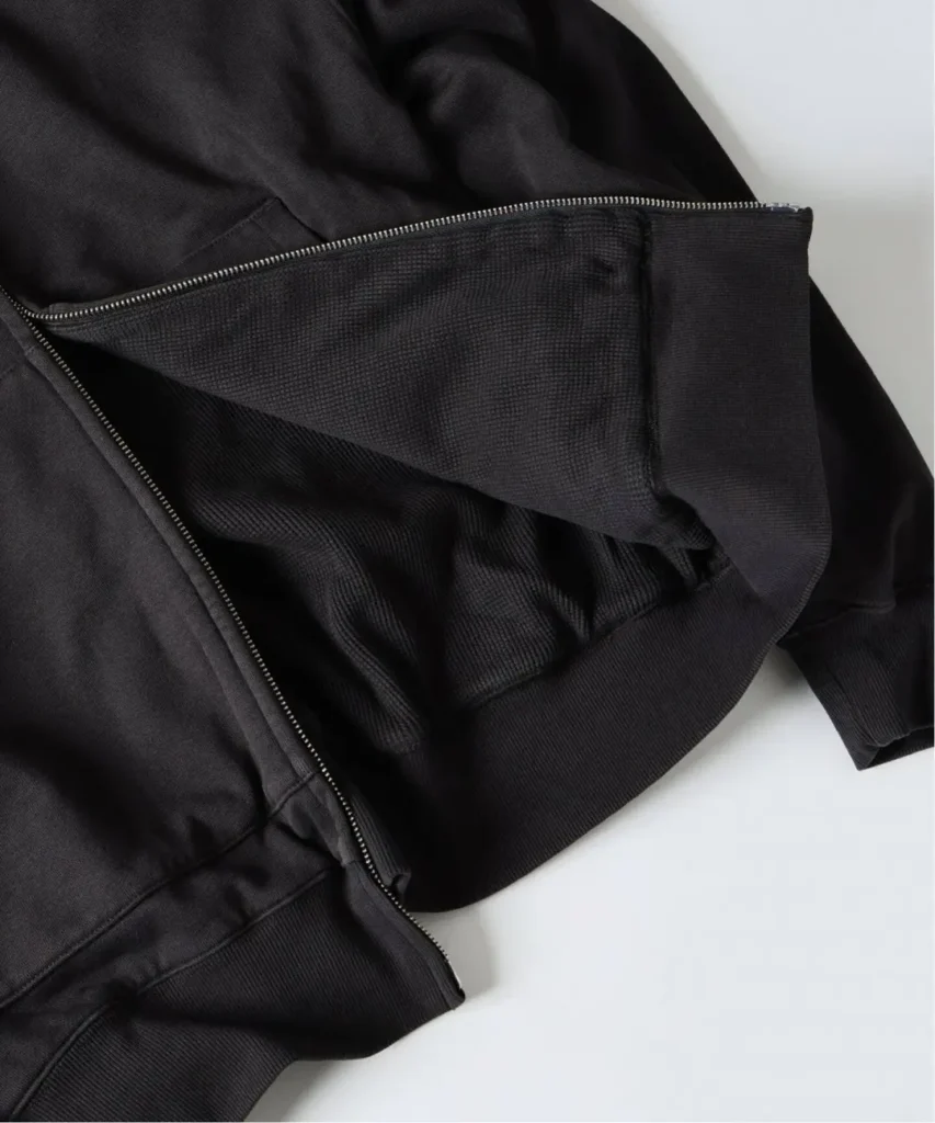Close-up of a black zip-up hoodie showing its textured inner layer, silver zipper and ribbed hem on a white surface