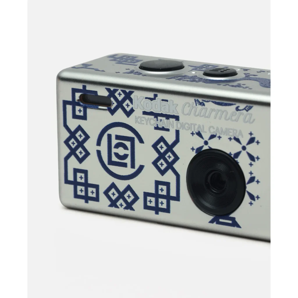 Close-up of a silver Kodak Charmera keychain digital camera with blue graphic patterns and a black lens on a white background