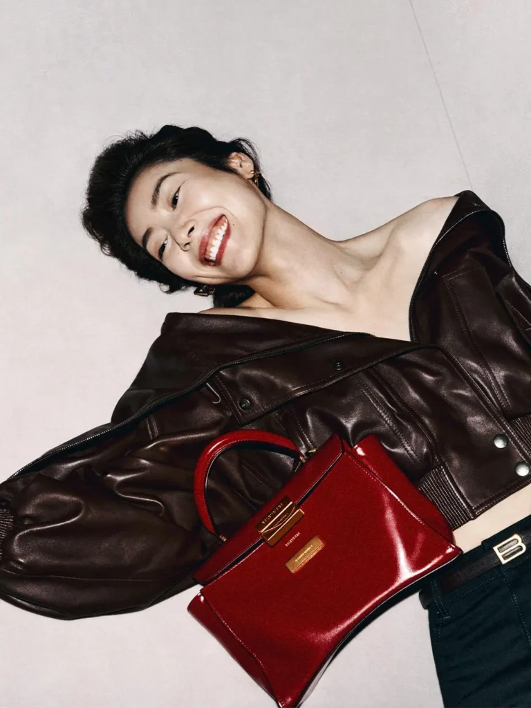 Smiling woman reclining in a dark brown leather jacket, with a structured red handbag across her torso against a pale