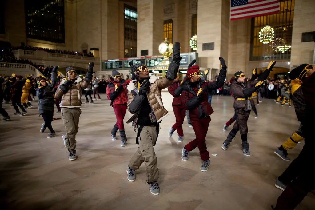 Dancers in padded winter clothing perform in Grand Central Terminal beneath chandeliers and an American flag