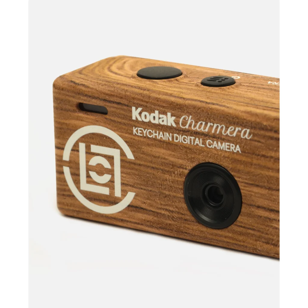 Close-up of a woodgrain Kodak Charmera keychain digital camera with black buttons, lens and white branding on a white